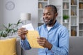 Senior joyful man sitting on sofa at home, satisfied smiling african american man reading mail message holding letter Royalty Free Stock Photo