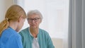 Senior lady patient with glasses talks to experienced young caretaker sitting near window in light room
