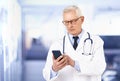 Senior male doctor text messaging on mobile phone while standing in doctor`s office Royalty Free Stock Photo