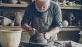 Senior potter is working at bottom part of ceramic pot on spinning potter& x27;s wheel in workplace. Creating traditional
