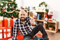 Senior man with beard wearing santa claus hat celebrating christmas at home, sitting on the floor by christmas tree and presents