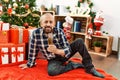 Senior man with beard wearing santa claus hat celebrating christmas at home, sitting on the floor by christmas tree and presents