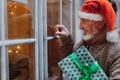 Senior man with a Christmas gift in hand standing in front of the window and knocking on it. Elderly man as Santa Claus Royalty Free Stock Photo