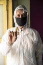 Senior man with corona virus preventive mask and PPE suit showing medicine ampul or vaccine in front of a window. Royalty Free Stock Photo