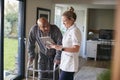 Senior Man In Dressing Gown Using Walking Frame Being Helped By Female Nurse With Digital Tablet