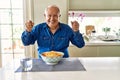 Senior man with grey hair eating pasta spaghetti at home very happy and excited doing winner gesture with arms raised, smiling and Royalty Free Stock Photo