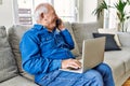 Senior man with grey hair sitting on the sofa at the living room of his house using computer laptop and speaking on the phone Royalty Free Stock Photo