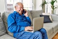 Senior man with grey hair sitting on the sofa at the living room of his house using computer laptop and speaking on the phone Royalty Free Stock Photo