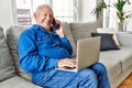 Senior man with grey hair sitting on the sofa at the living room of his house using computer laptop and speaking on the phone Royalty Free Stock Photo