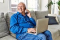 Senior man with grey hair sitting on the sofa at the living room of his house using computer laptop and speaking on the phone Royalty Free Stock Photo