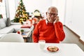 Senior man with grey hair sitting on the table with cookies by christmas tree looking stressed and nervous with hands on mouth