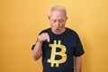 Senior man with grey hair wearing bitcoin t shirt pointing down with fingers showing advertisement, surprised face and open mouth