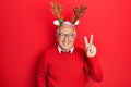 Senior man with grey hair wearing deer christmas hat smiling with happy face winking at the camera doing victory sign