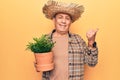 Senior man with grey hair wearing gardener hat holding plant pot pointing thumb up to the side smiling happy with open mouth Royalty Free Stock Photo