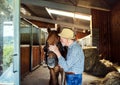 A senior man with a hat standing close to a horse in a stable, holding it. Royalty Free Stock Photo