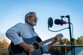 Senior man imposing musician plays the guitar and sings into microphone. Elegant elderly male 60 bearded performs outdoors in