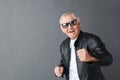 Senior man in leather jacket and sunglasses standing isolated on gray looking camera dancing cheerful