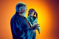 Senior man and little girl, grandfather and granddaughter watching TV over gradient orange background in neon light