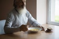 Old sad man with a long gray beard sitting by the table and eating soup and bread Royalty Free Stock Photo