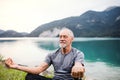 A senior man pensioner sitting by lake in nature, doing yoga exercise.