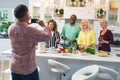 Senior man photographing smiling multiracial male and female friends preparing food at home