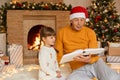 Senior man reading book to granddaughter at Christmas Eve, family sitting on floor near fireplace and xmas tree, female kid with Royalty Free Stock Photo