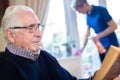 Senior Man Reading In Foreground Whilst Female Home Help Cleans House