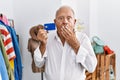 Senior man at retail shop holding credit card covering mouth with hand, shocked and afraid for mistake