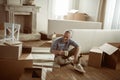 Senior man sitting on floor with cup of tea and taking break from packing cardboard boxes, moving out concept