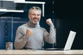 A senior man is sitting in the office Celebrates victory, success, shows strength with his hands Royalty Free Stock Photo