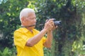 Senior man taking a photo by a digital camera while standing in the park. An elderly Asian man wears a yellow shirt be happy when