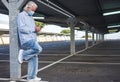 A senior man wearing medical mask due to coronavirus standing under the metal structure of a deserted parking looking at his smart