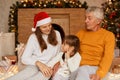 Senior man wearing orange sweater sitting with his family near xmas tree indoor, people spending new year eve together, enjoying Royalty Free Stock Photo