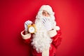 Senior man wearing Santa Claus costume holding alarm clock over isolated red background cover mouth with hand shocked with shame Royalty Free Stock Photo