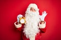 Senior man wearing Santa Claus costume holding alarm clock over isolated red background very happy and excited, winner expression Royalty Free Stock Photo