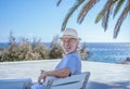 Senior man wearing straw cap sitting outdoor at sea looking at camera smiling - horizon over water Royalty Free Stock Photo