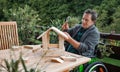 Senior man in wheelchair constructing birdhouse outdoors on terrace, diy project.