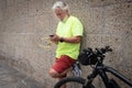 Senior man with white hair and beard, looking at his cell phone near his bicycle. Brick wall as a background. Dressed in a Royalty Free Stock Photo