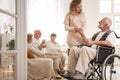 Senior man on wheelchair with helpful nurse holding his hand and friends sitting on couch drinking tea Royalty Free Stock Photo