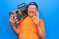 Senior modern handsome grey-haired man wearing cap listening to music holding boombox doing ok sign with fingers, smiling friendly