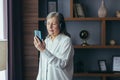 Senior modern woman at home by the window listens to music on the phone, uses a music application for online listening Royalty Free Stock Photo