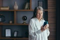 Senior modern woman at home by the window listens to music on the phone, uses a music application for online listening, and Royalty Free Stock Photo