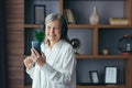 Senior modern woman at home by the window listens to music on the phone, uses a music application for online listening, and Royalty Free Stock Photo