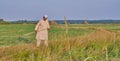 An elderly Muslim man in an embroidered skullcap and white traditional Clothes mows hand-scythe grass in a hayfield