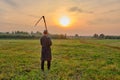 An senior Muslim man in a skullcap and traditional clothes leaves with a hand scythe from the hay field at sunset