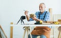 Senior old Caucasian man wearing check shirt, apron, making DIY wooden furniture, using mobile phone, streaming live video clip, Royalty Free Stock Photo