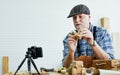 Senior old Caucasian man wearing check shirt, apron, making DIY wooden furniture, using mobile phone, streaming live video clip, Royalty Free Stock Photo