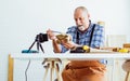 Senior old Caucasian man wearing check shirt, apron, making DIY wooden furniture, using mobile phone, streaming live video clip, Royalty Free Stock Photo