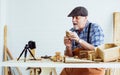 Senior old Caucasian man wearing check shirt, apron, making DIY wooden furniture, using mobile phone, streaming live video clip, Royalty Free Stock Photo