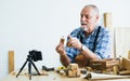 Senior old Caucasian man wearing check shirt, apron, making DIY wooden furniture, using mobile phone, streaming live video clip, Royalty Free Stock Photo